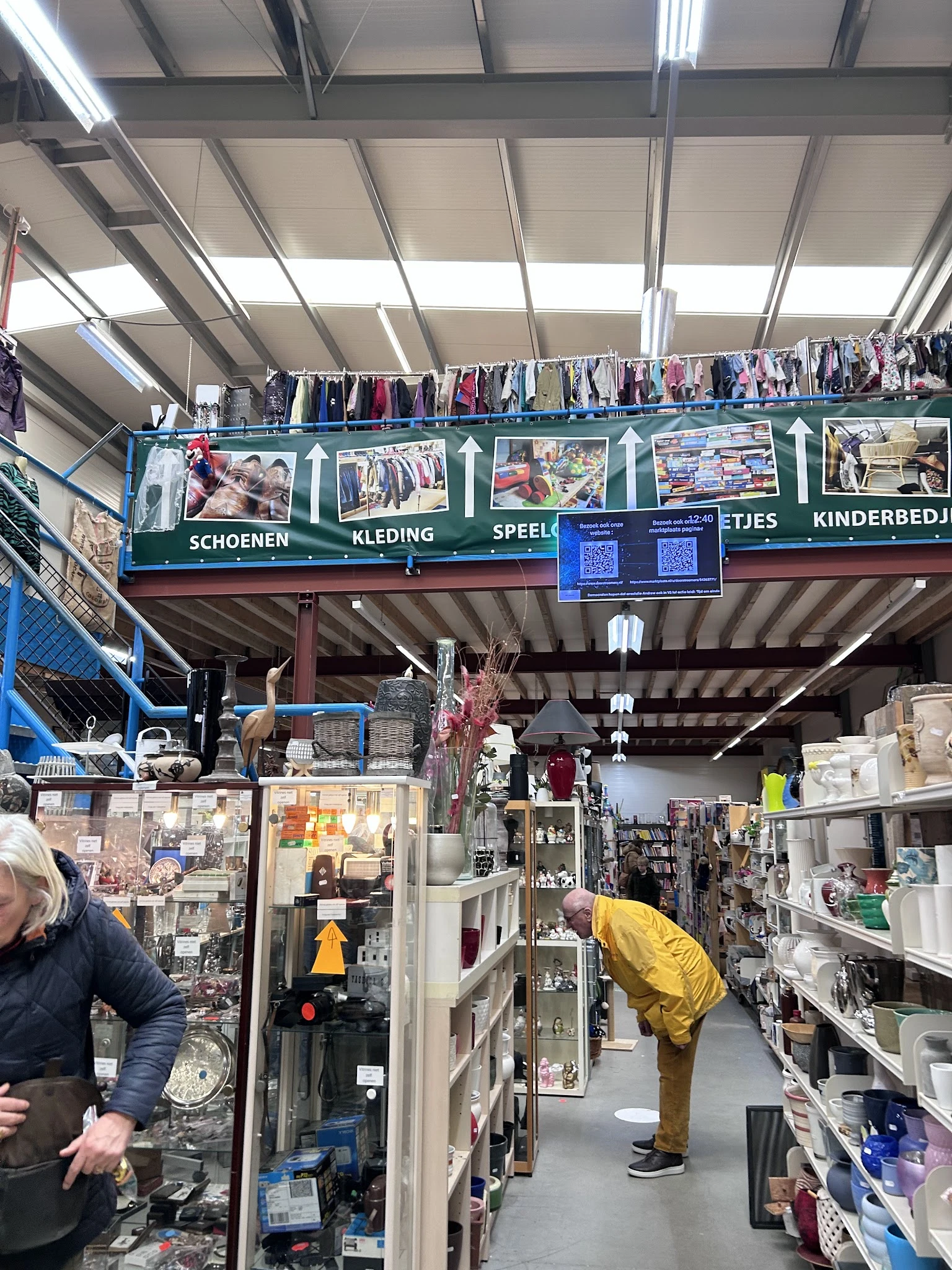 Overdekt winkelinterieur met kleding aan rekken en aan het plafond, en diverse spullen op tafels en stellingen.