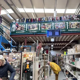Overdekt winkelinterieur met kleding aan rekken en aan het plafond, en diverse spullen op tafels en stellingen.