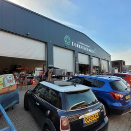 Voorkant van kringloopwinkel met grijze gevel, grote witte garagedeuren en parkeerplaats met auto's