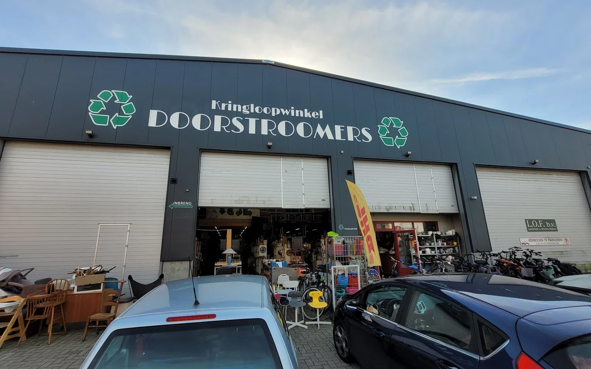 Gevel van kringloopwinkel met drie open deuren, uitgestalde spullen en voertuigen ervoor
