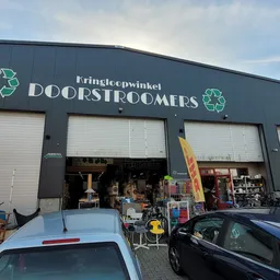 Gevel van kringloopwinkel met drie open deuren, uitgestalde spullen en voertuigen ervoor