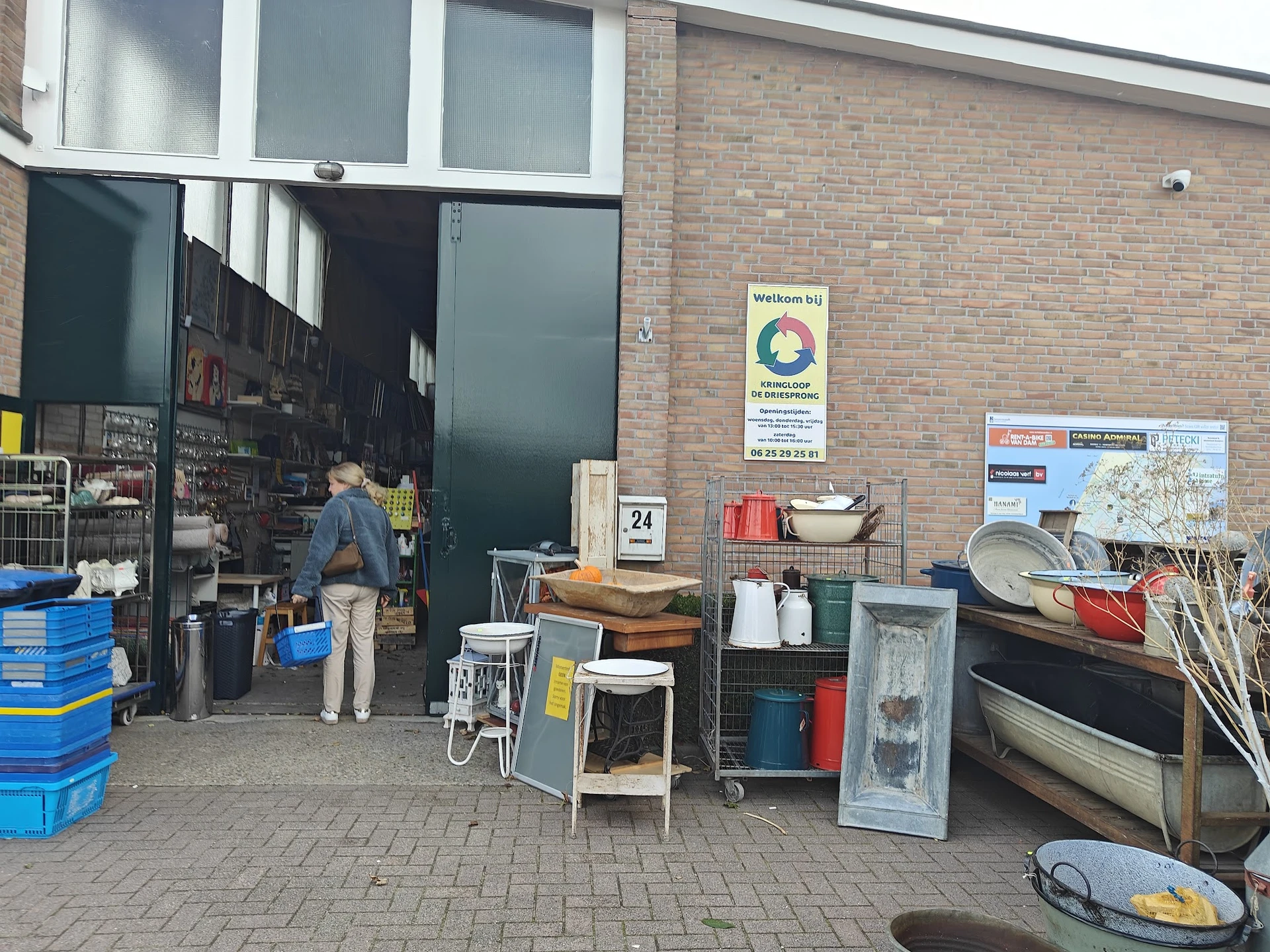 Ingang van kringloopwinkel met open deur, spullen op straat en man in deuropening