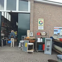 Ingang van kringloopwinkel met open deur, spullen op straat en man in deuropening