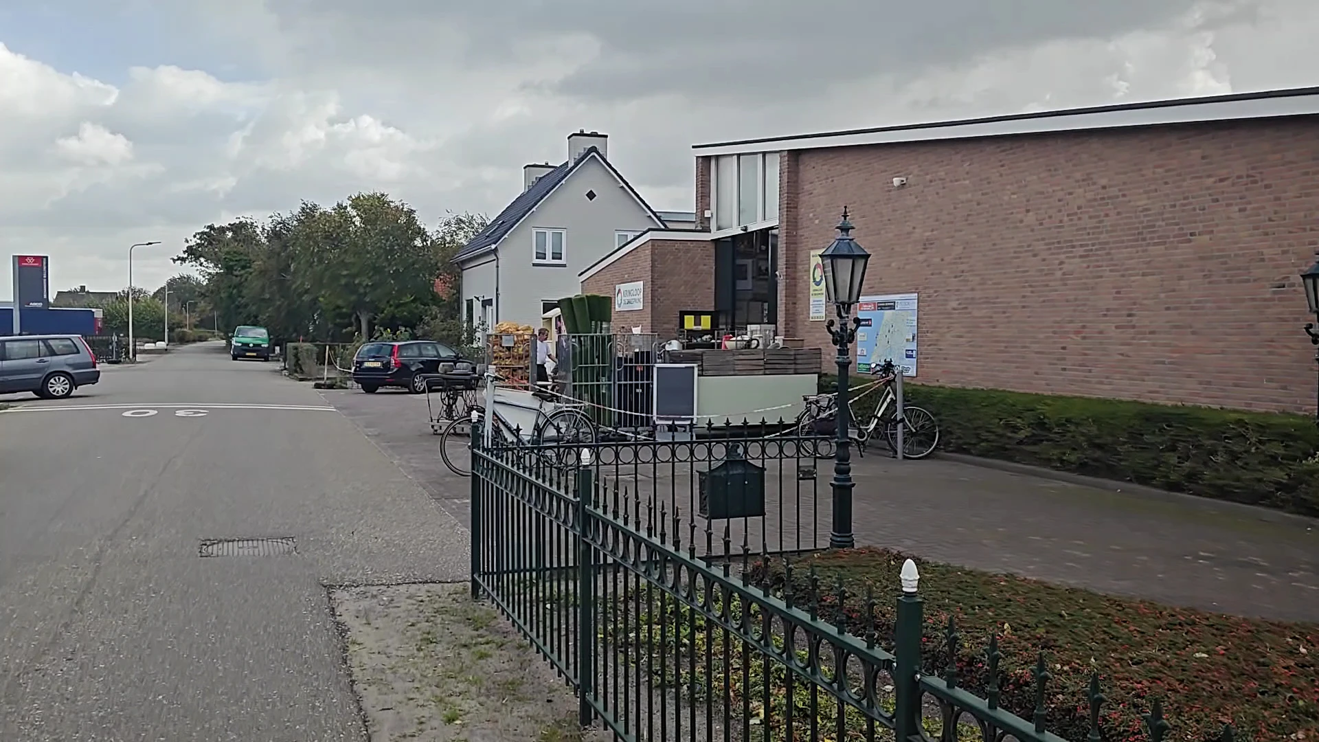 Voorkant van kringloopwinkel met bakstenen gevel, zwart hek en enkele fietsen geparkeerd