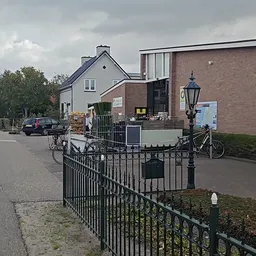 Voorkant van kringloopwinkel met bakstenen gevel, zwart hek en enkele fietsen geparkeerd