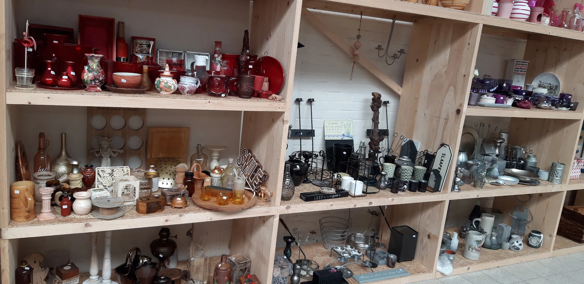Houten stellingkasten met verzameling tweedehands huishoudartikelen, waaronder rode en zwarte kaarsen, flessen, elektronische