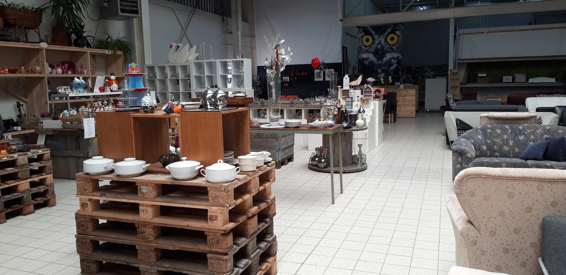 Ruime winkelruimte met diverse uitgestalde spullen op houten pallets en vitrines, witte tegelvloer