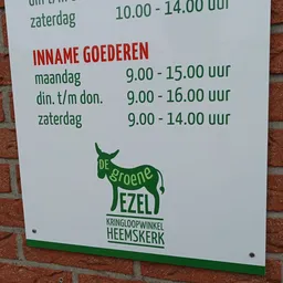 Openingstijden van kringloopwinkel De Groene Ezel op een wit bord tegen een bakstenen muur