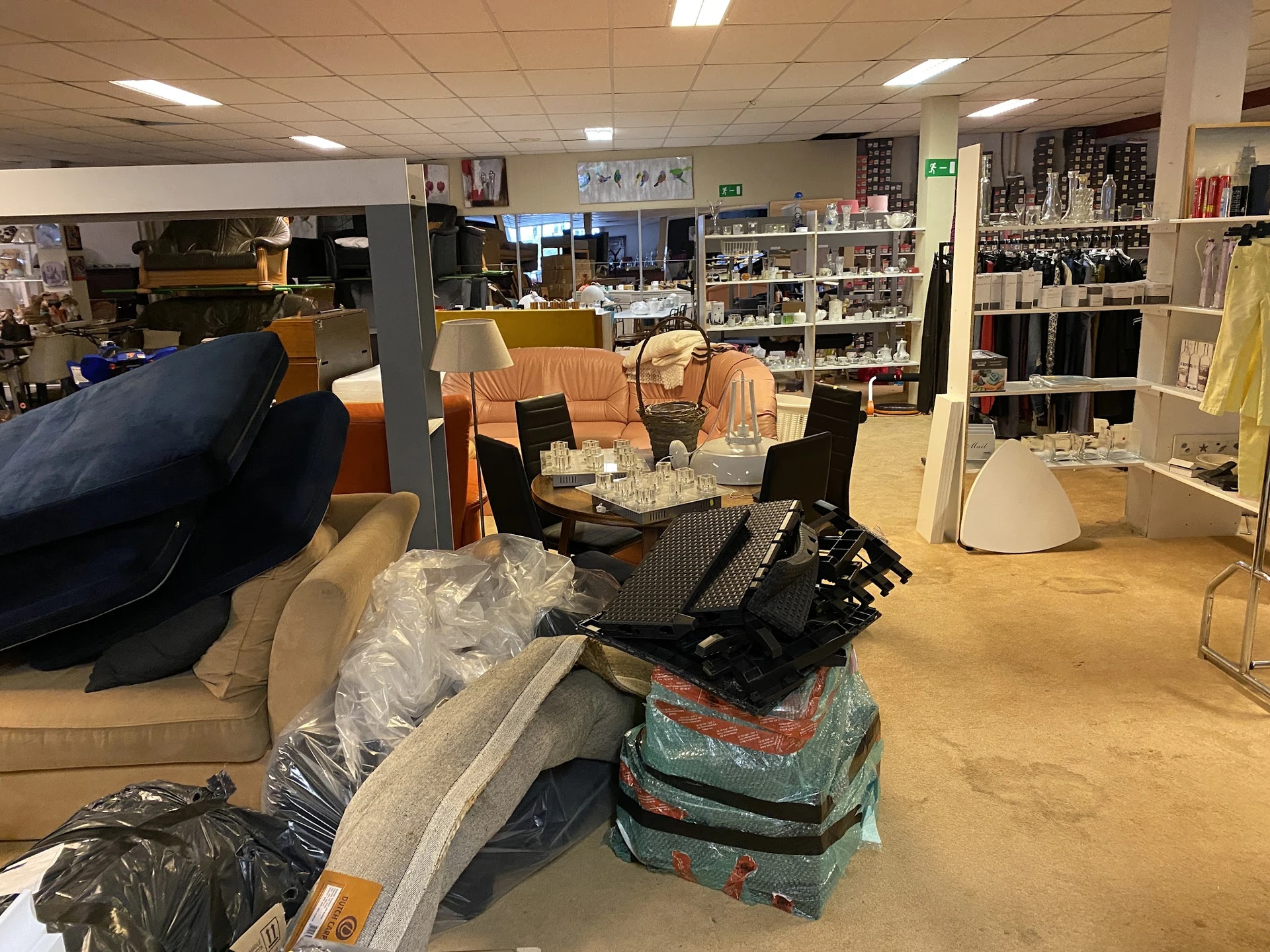 Winkelvloer met diverse meubels en dozen gestapeld, stellingen met spullen op de achtergrond