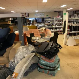 Winkelvloer met diverse meubels en dozen gestapeld, stellingen met spullen op de achtergrond