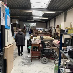 Winkelvloer van kringloopwinkel met meubels, tafels, stoelen en diverse spullen uitgestald onder fluorescente verlichting