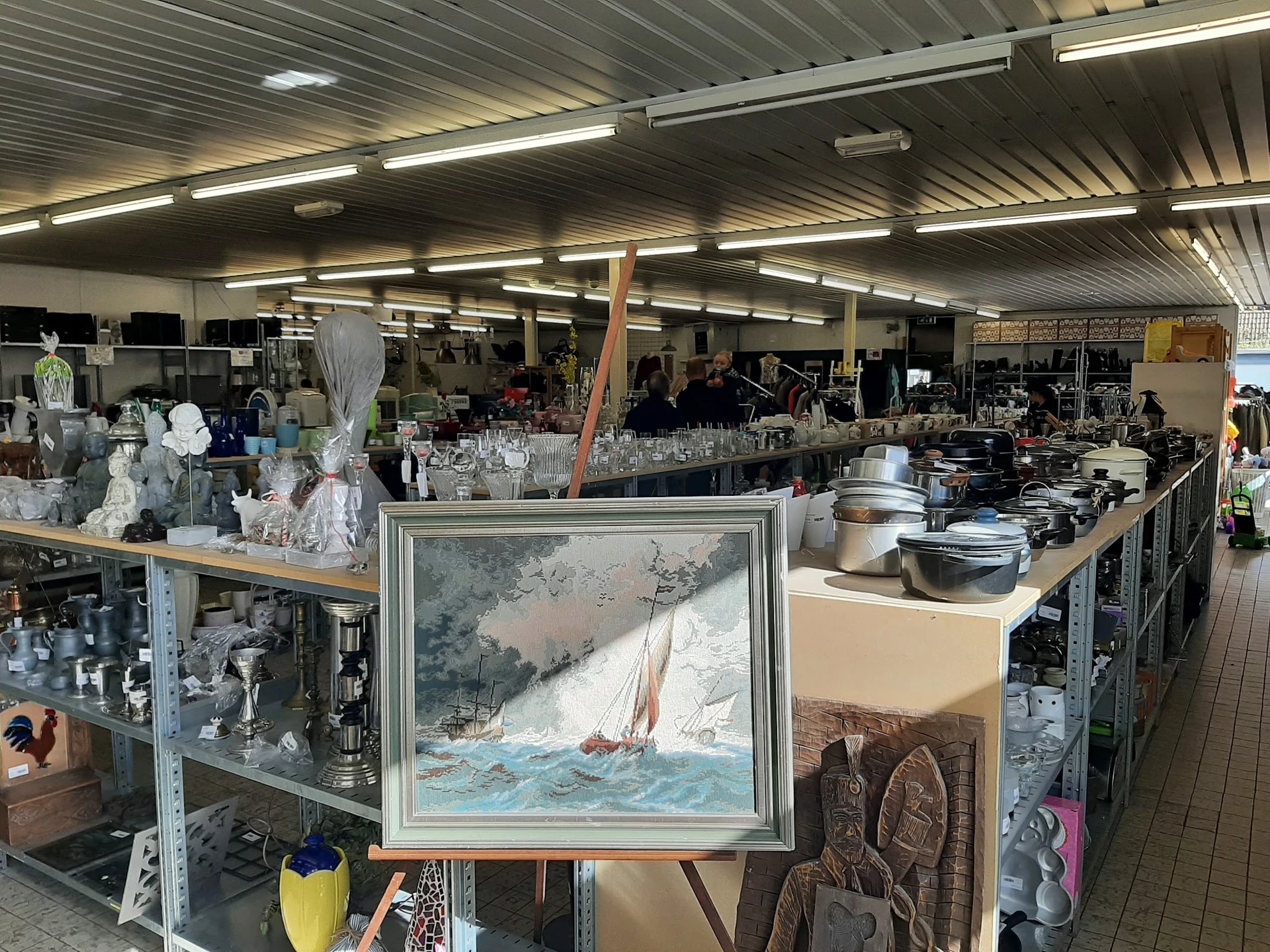 Overzicht van de kringloopwinkel met stellingen vol spullen, een schilderij op een ezel vooraan, en diverse huishoudelijke ar