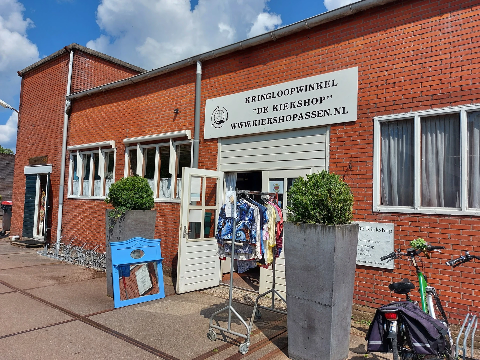 Rode bakstenen gevel van kringloopwinkel met witte deuren, uithangbord en fietsen ervoor