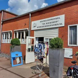 Rode bakstenen gevel van kringloopwinkel met witte deuren, uithangbord en fietsen ervoor