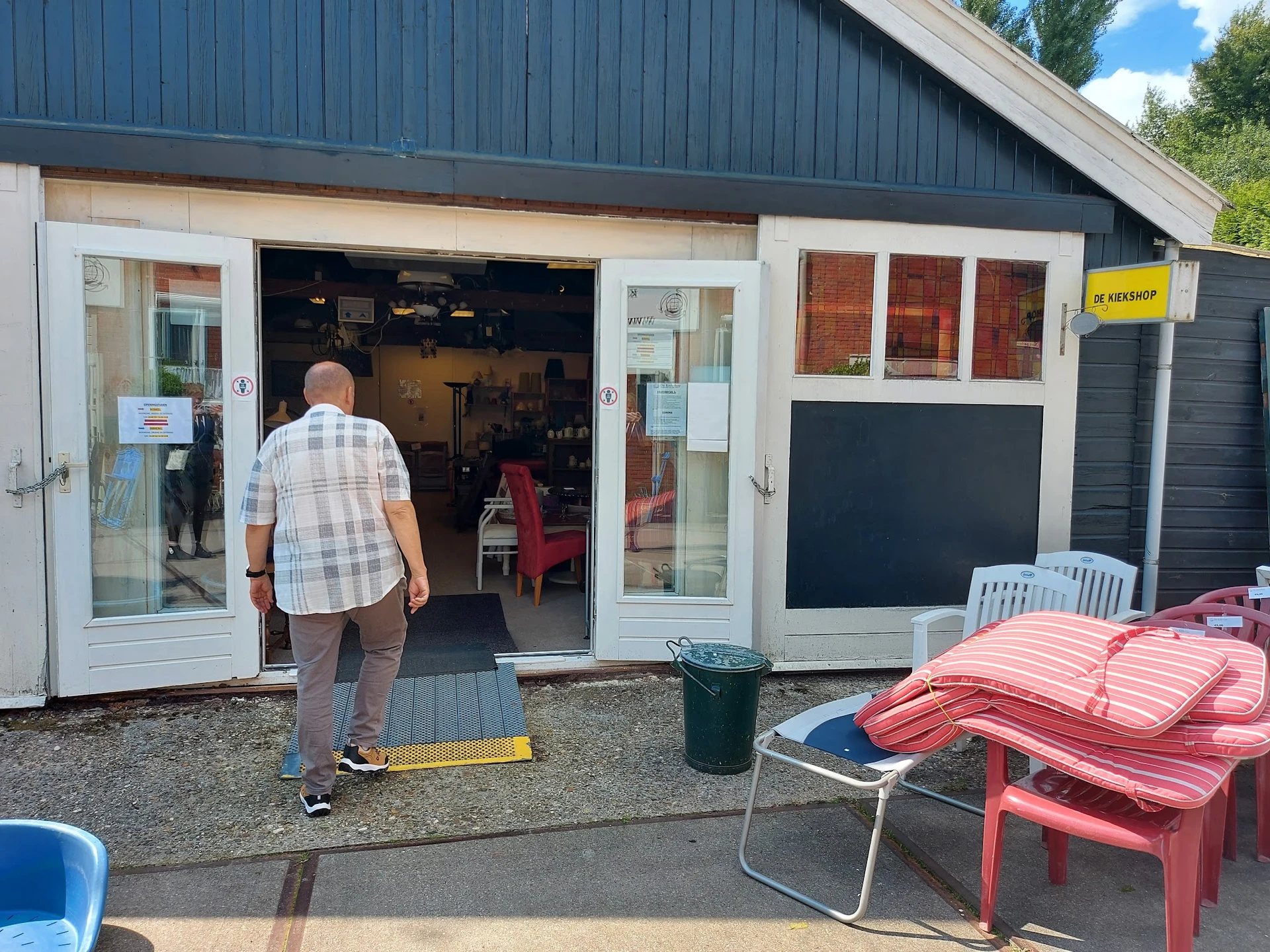 Voorgevel van kringloopwinkel met open deuren, terras met stoelen en tuinmeubilair ervoor
