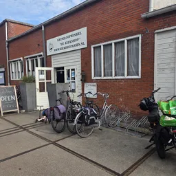 Voorgevel van kringloopwinkel met rode bakstenen, ingang met winkelnaam boven deur, fietsen geparkeerd ervoor