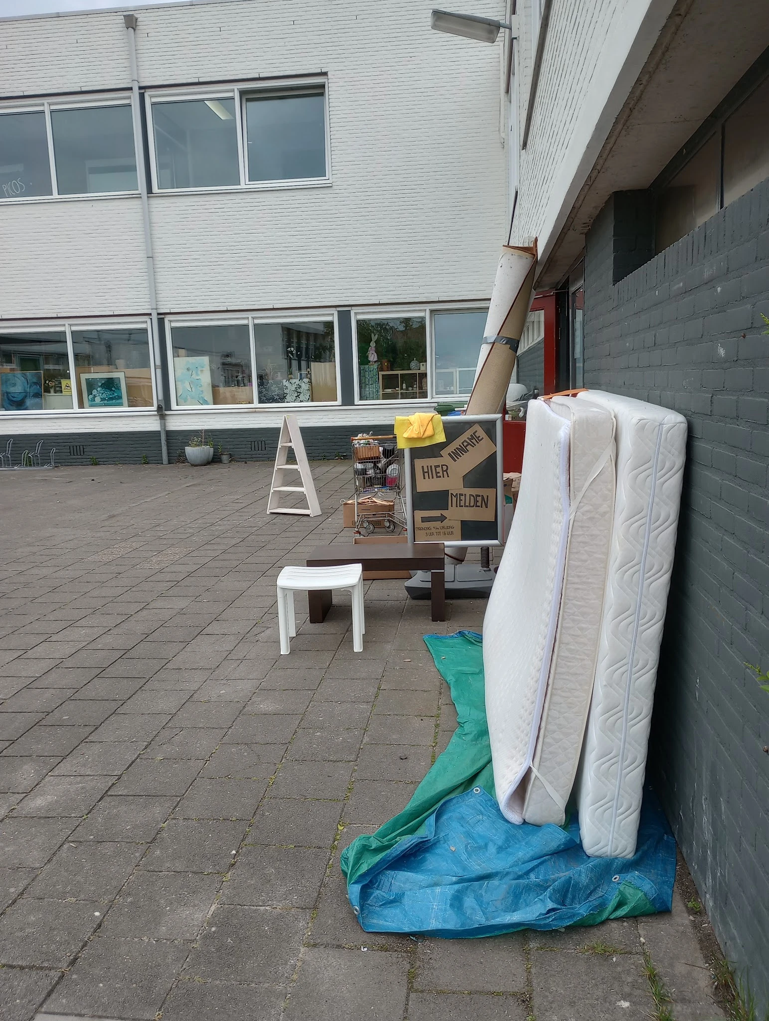 Tweedehands meubels buiten uitgestald: matrassen, tafels en stoelen op het terras van de winkel