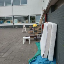 Tweedehands meubels buiten uitgestald: matrassen, tafels en stoelen op het terras van de winkel