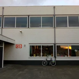 Voorgevel van kringloopwinkel met witte bakstenen muur, grote ramen en rode letters 'GD' op de deur