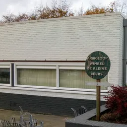 Witte gevel van kringloopwinkel met grote ramen en groen uithangbord met winkelnaam