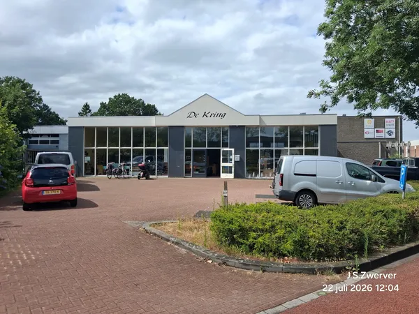 Grijze gevel van kringloopwinkel De kring met grote ramen, parkeerplaats met auto's en groene struiken