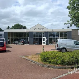 Grijze gevel van kringloopwinkel De kring met grote ramen, parkeerplaats met auto's en groene struiken