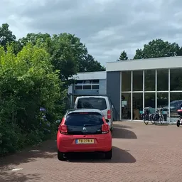 Parkeerplaats voor de kringloopwinkel met rode auto en voetgangers op bakstenen oppervlak