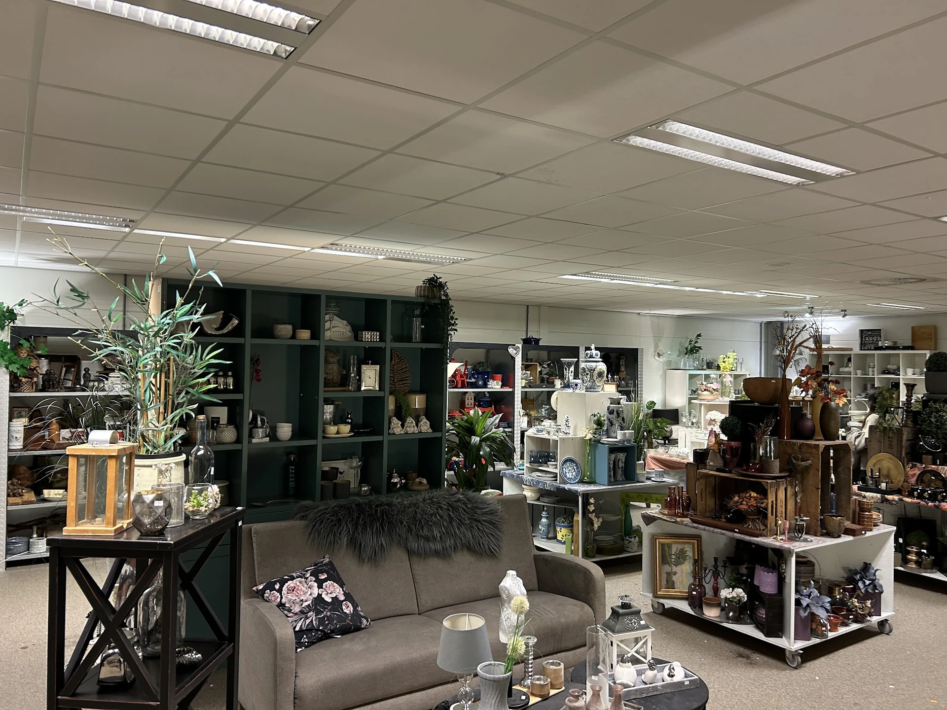 Winkelvloer met stellingen vol huisraad, decoratieartikelen en een grijze bank als display in kringloopwinkel