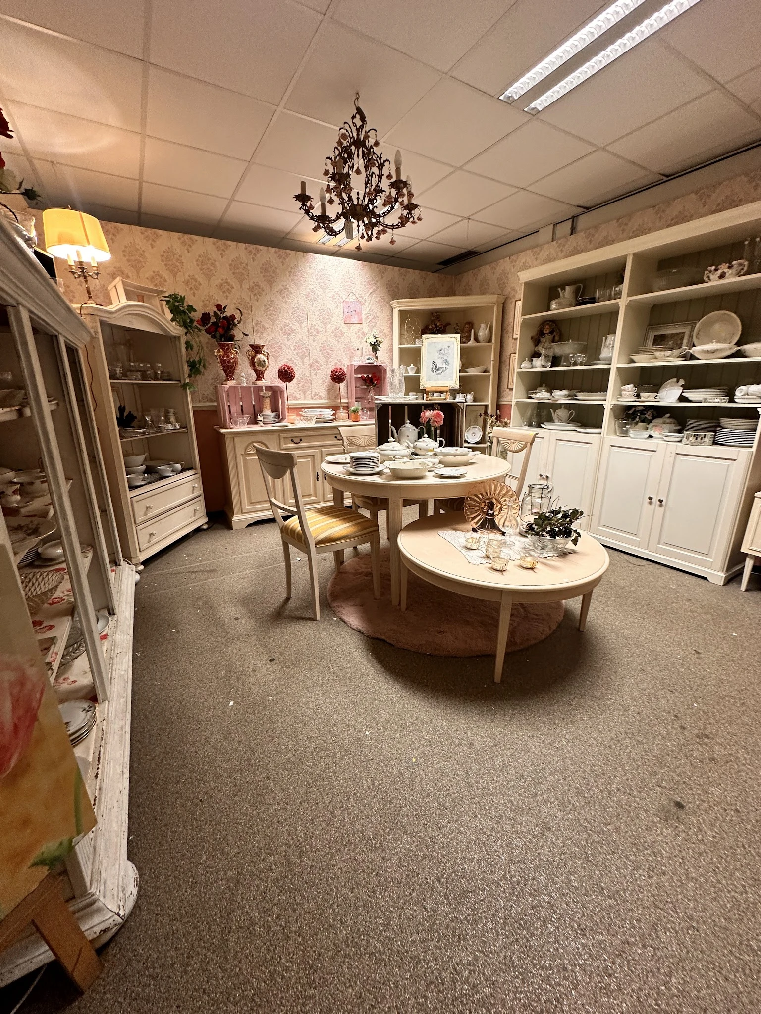 Winkelinterieur met witte kasten, twee ronde tafels met servies, roze wandafwerking en decoratieve items.