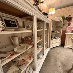 Witte kasten met glasdeuren vol serviesgoed, borden en decoratieve voorwerpen in een kringloopwinkel