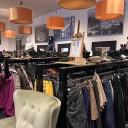 Rekken met tweedehands kleding, inclusief jassen, broeken en accessoires onder koperen lampen