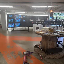 Winkelvloer met oranje markering, stellingen met uitgestalde spullen, displays met monitoren aan de muur, en een houten tafel
