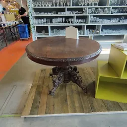 Ronde houten tafel met ornamentaal gesneden voet en decoratieve rand, staande in een kringloopwinkel
