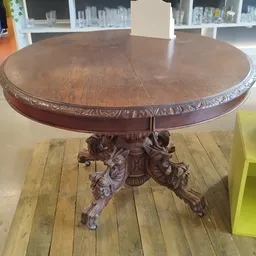 Ronde houten tafel met ornamentaal gesneden voet en decoratieve rand, geplaatst op gele vloerplanken