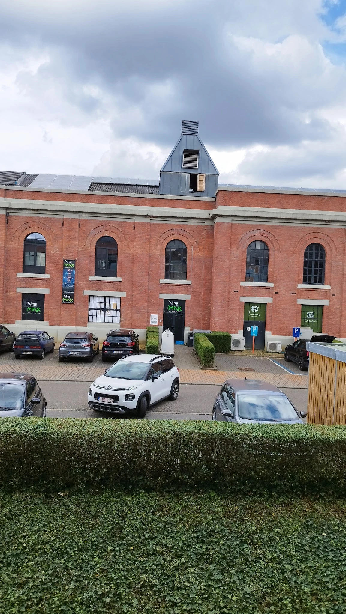 Bakstenen gevel van kringloopwinkel met groene deuren, parkeerplaats met auto's voor het pand