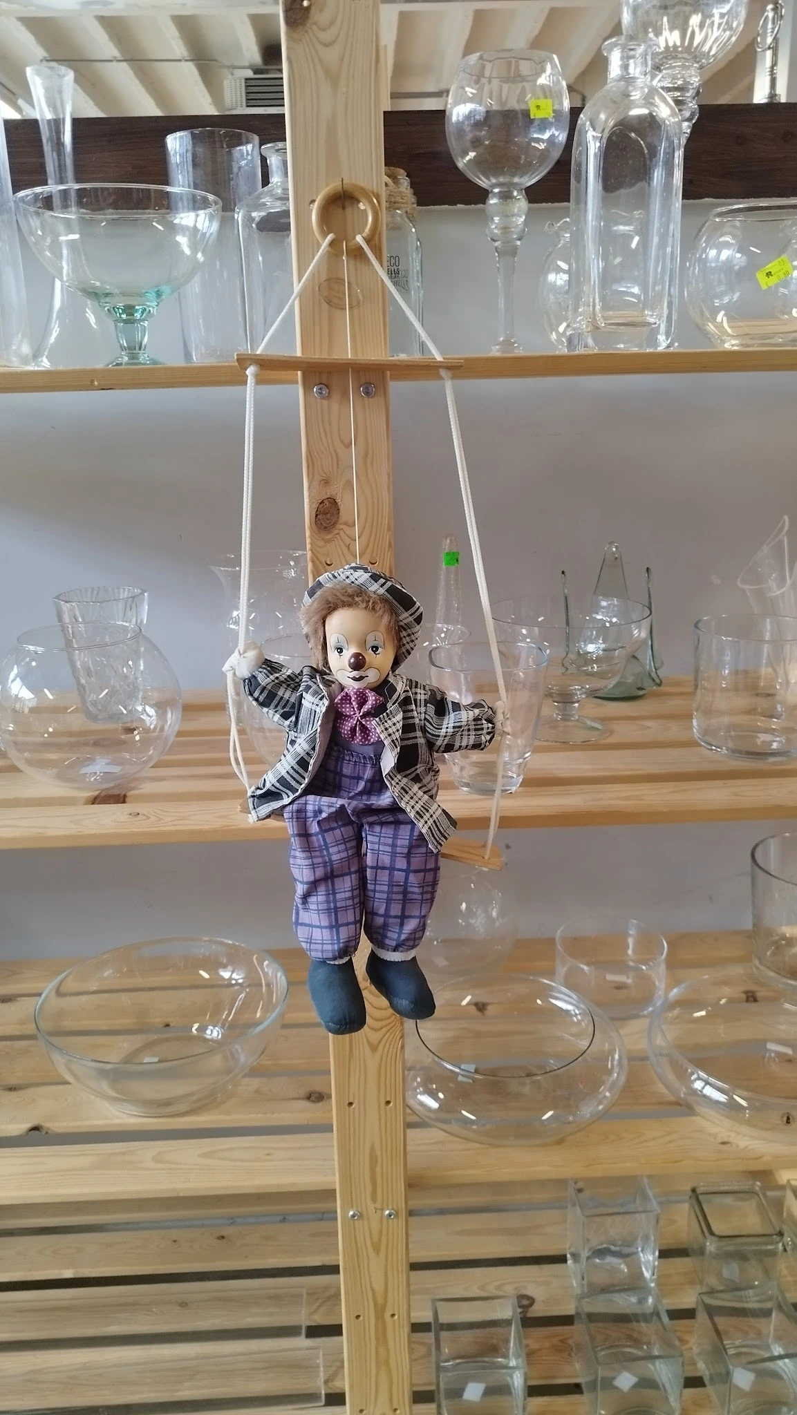 Pop met geruit jasje en paarse broek, hangend aan touwen boven een houten tafel met glazen