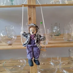 Pop met geruit jasje en paarse broek, hangend aan touwen boven een houten tafel met glazen