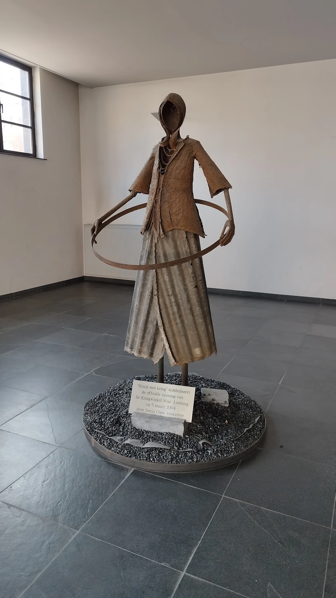 Bronzen beeldhouwwerk van een figuur met uitgestrekte armen op een stenen sokkel met naamplaatje