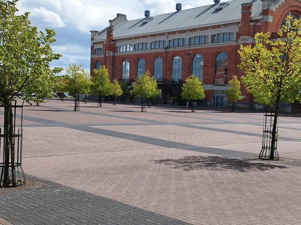 Rood bakstenen gebouw met grote ramen aan een plein met tegels en jonge bomen
