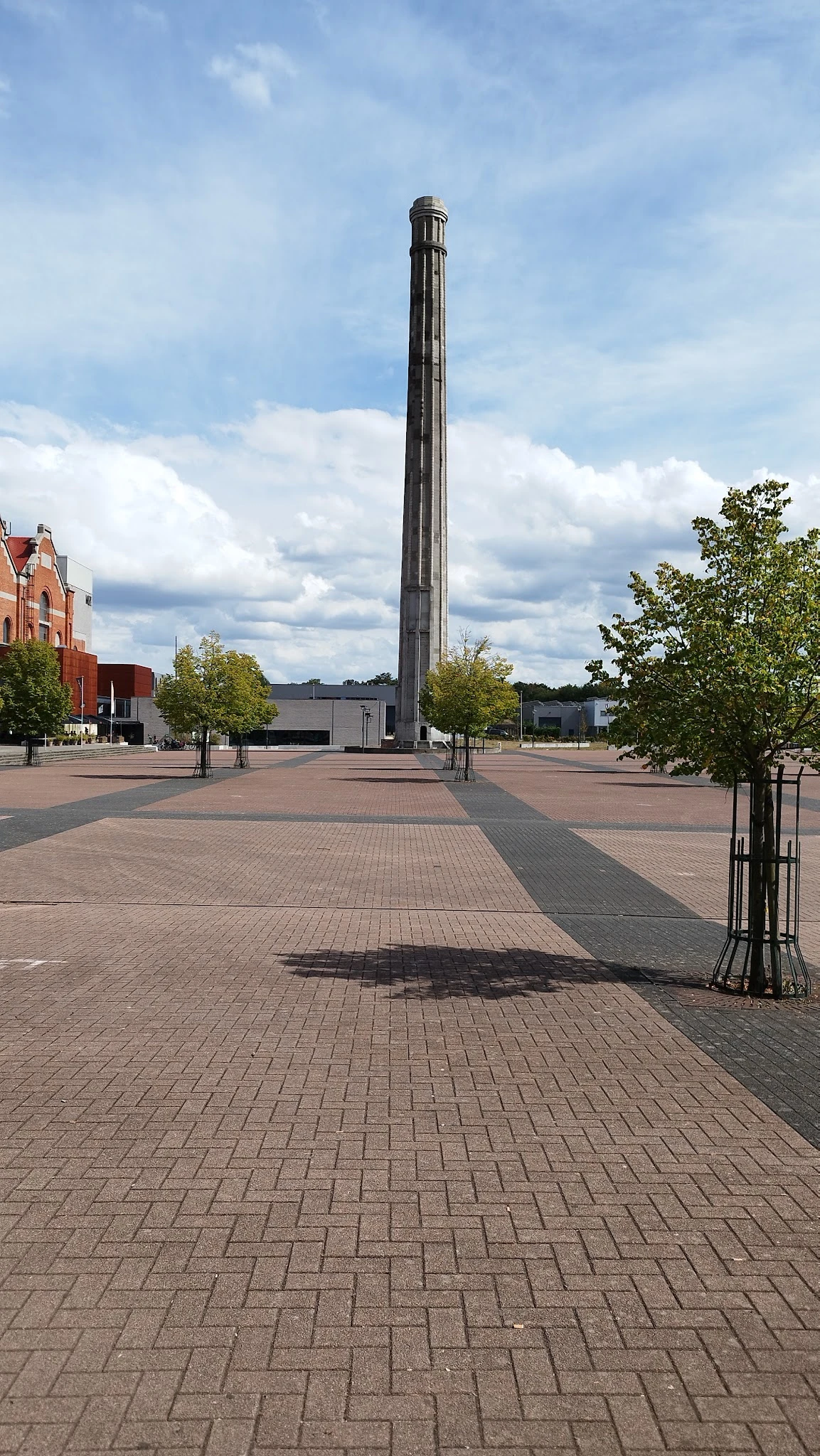 Groot plein met tegel bestrating, hoge zuil in het midden, rode bakstenen gebouw links, enkele jonge bomen