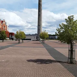 Groot plein met tegel bestrating, hoge zuil in het midden, rode bakstenen gebouw links, enkele jonge bomen