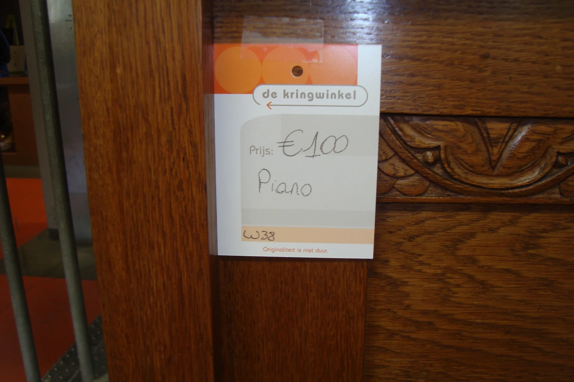 Prijskaartje van 'de kringwinkel' op houten meubelstuk met aanduiding 'Piano' en prijs €100