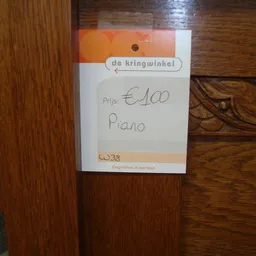 Prijskaartje van 'de kringwinkel' op houten meubelstuk met aanduiding 'Piano' en prijs €100