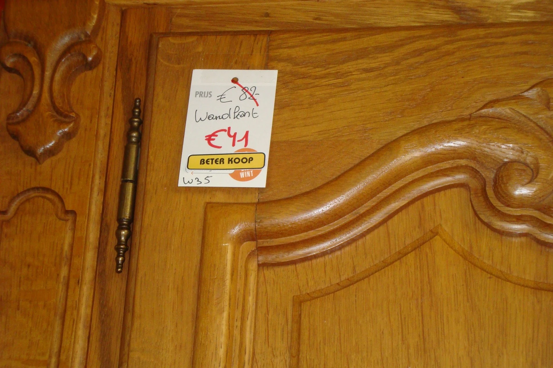 Houten meubeldeel met golvende carving en prijssticker, naast een penseel op houten ondergrond