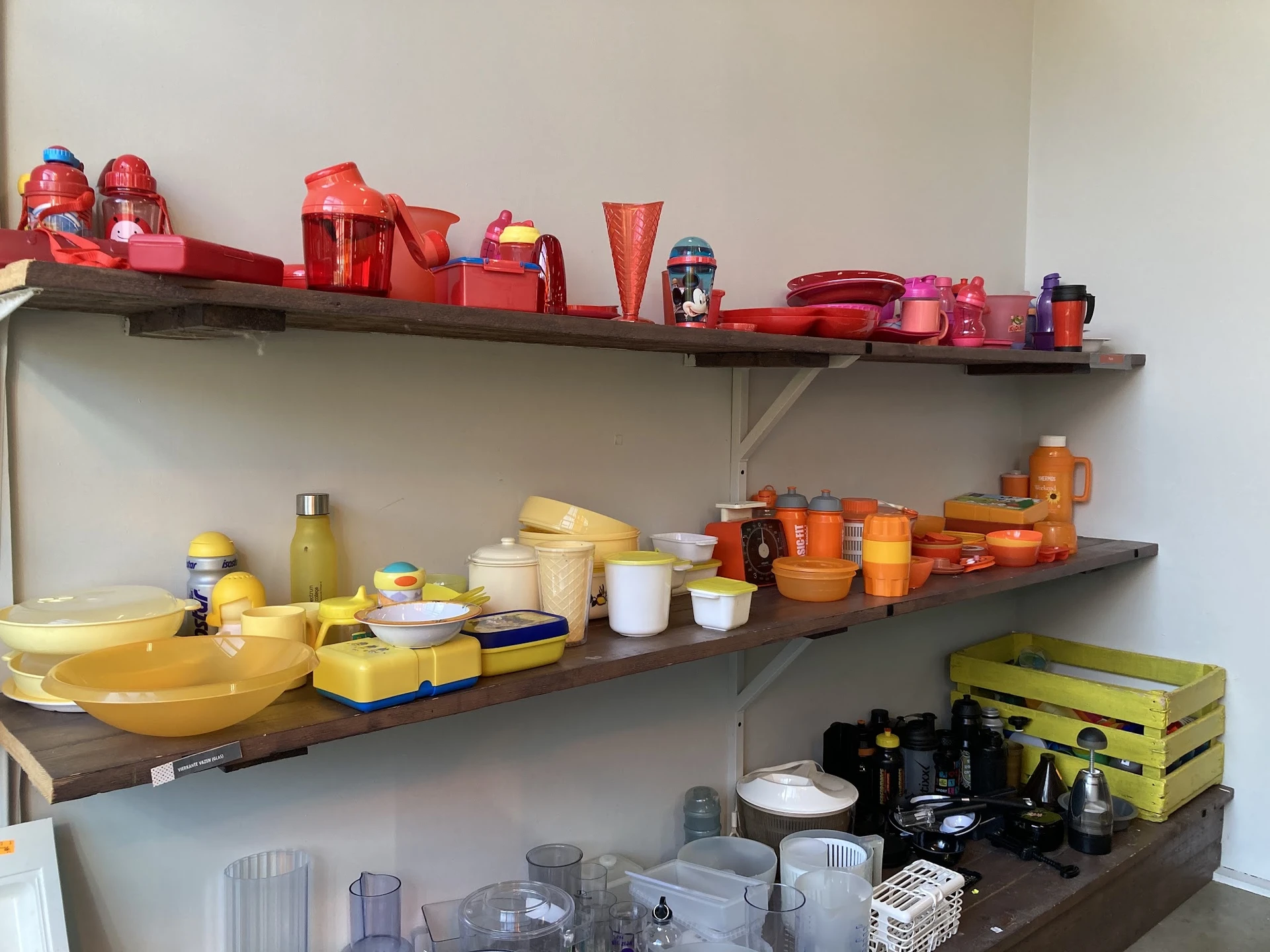 Drie wandplanken met huishoudelijke items: rode keukenaccessoires op de bovenste plank, gele servies en bestek op de middelst