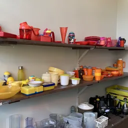 Drie wandplanken met huishoudelijke items: rode keukenaccessoires op de bovenste plank, gele servies en bestek op de middelst