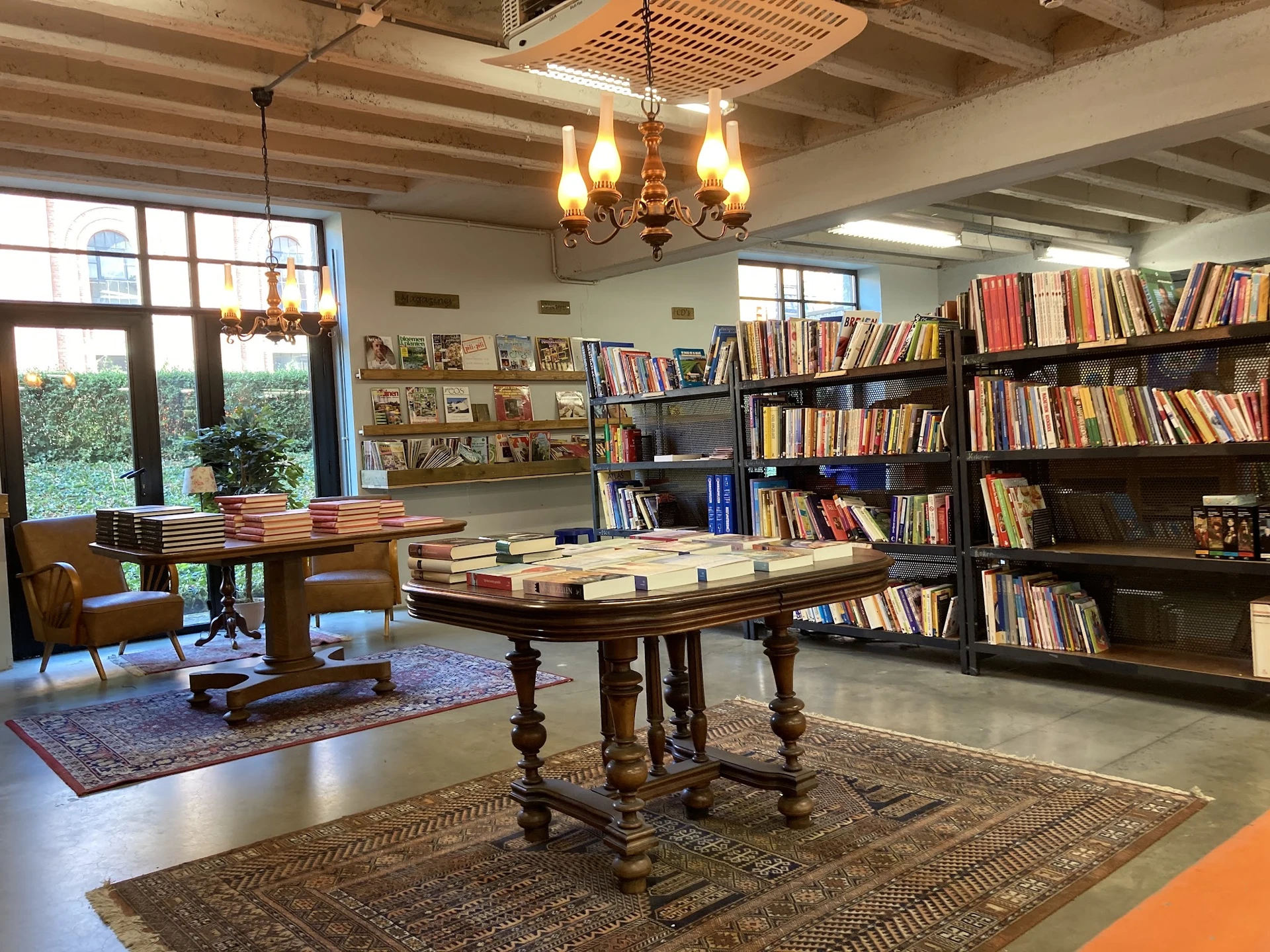 Ruime winkelruimte met boekenkasten, houten tafels met boeken, kroonluchter en grote ramen met uitzicht op groen.