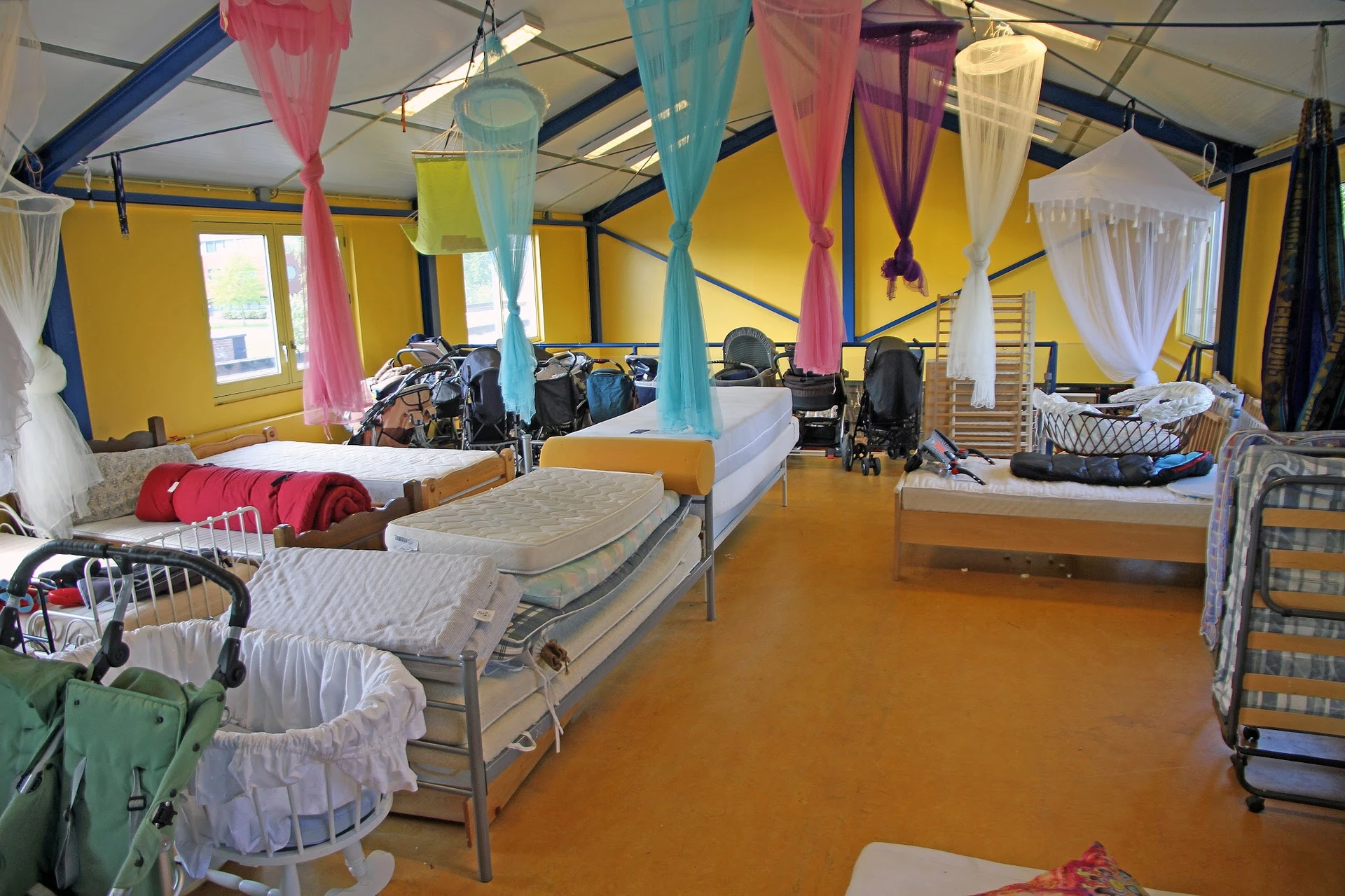 Slaapzaal met meerdere bedden, gekleurde doeken aan het plafond en opgestelde beddenspullen