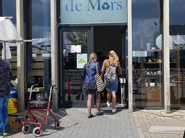 Ingang van kringloopwinkel de Mors met twee vrouwen in de deuropening, winkelnaam zichtbaar boven
