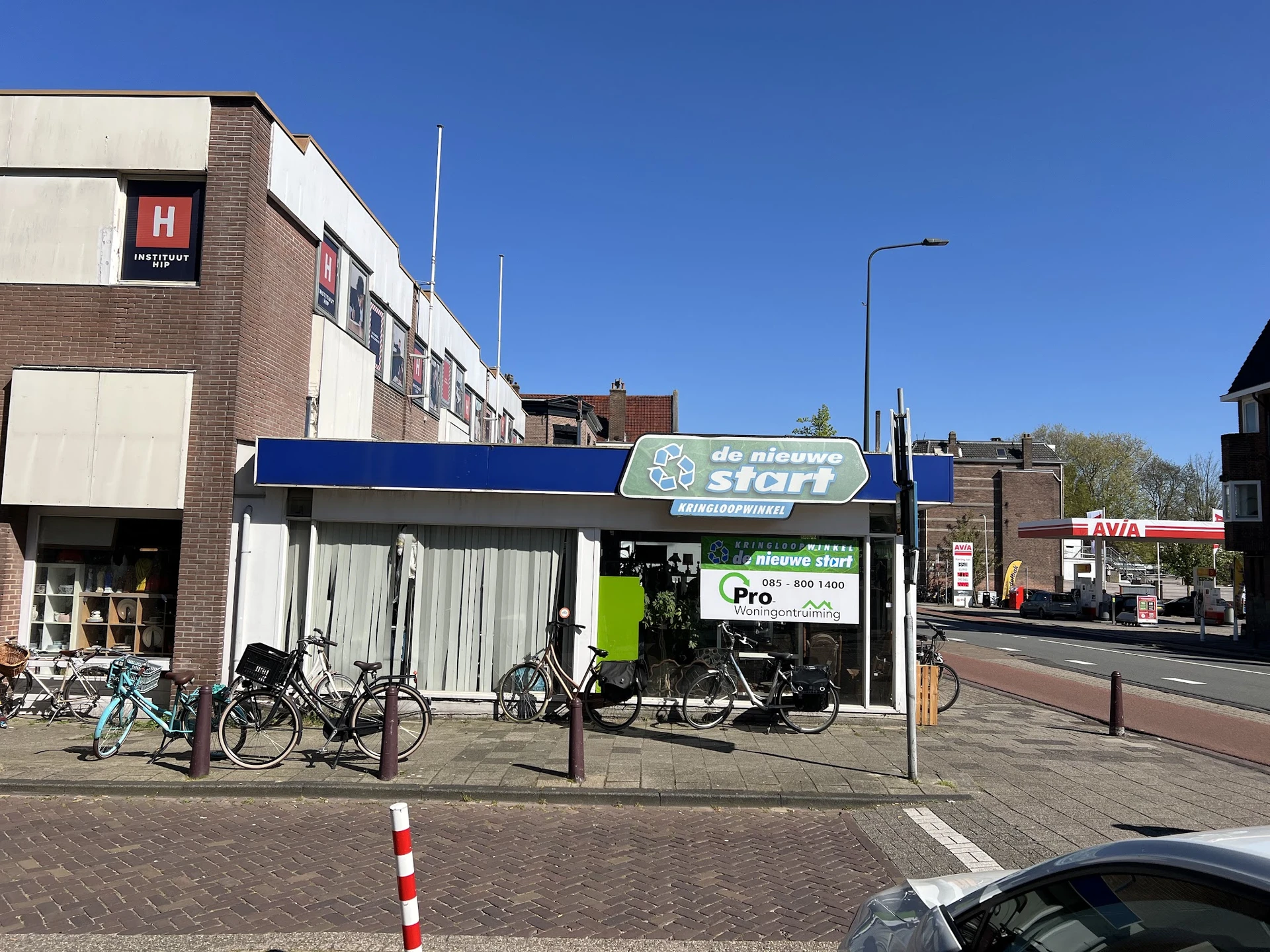 Gevel van kringloopwinkel De Nieuwe Start met blauwe luifel, fietsen geparkeerd ervoor en helder weer